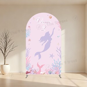 May include: An arched backdrop display featuring a light pink background with a purple mermaid silhouette, the text "Vernus" and "2", and various illustrations of coral, starfish, and seashells along the bottom and sides of the panel.