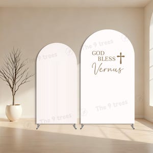 May include: Two white arched backdrops. One has a textured, vertical striped design. The other has the text "GOD BLESS Vernus" with a cross symbol. Both backdrops have metal stands and are for events.