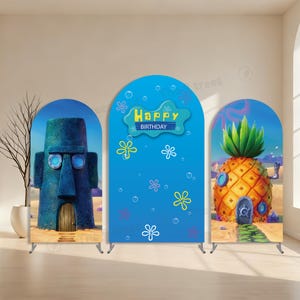 Può includere: Tre espositori di sfondo ad arco con immagini di cartoni animati. Uno raffigura una statua di pietra blu con una porta, un altro ha uno sfondo blu con il testo "Happy Birthday", e l'ultimo mostra una casa ananas. Il tutto è ambientato su una spiaggia sabbiosa.
