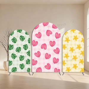 May include: Three arched backdrop stands featuring fabric covers with distinct patterns. The left stand has green monstera leaves on a white plaid background. The center stand displays pink anthurium flowers on a pink plaid. The right stand shows yellow plumeria flowers on a yellow plaid.