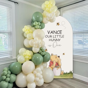 May include: A party decoration with a Winnie the Pooh theme. The design includes a large sign with the text "Vance Our Little Hunny is One" and a cartoon illustration of Winnie the Pooh. The sign is surrounded by a balloon arch in shades of green, white, and yellow.