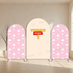 Op de afbeelding: Drie gebogen achtergrondstandaards. Twee roze stands hebben een wit wolkenpatroon. De middelste standaard is beige met de tekst "VERNUS STORY 1" in geel en rood. De stands zijn voor een feest of evenement.