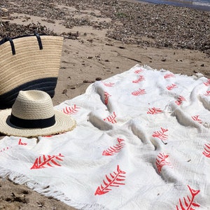 Op de afbeelding: Wit strandkleed met een rood visgraatpatroon. Het kleed is gevouwen en heeft een gefranst randje.