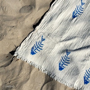 Op de afbeelding: Een witte strandhanddoek met een blauw visgraatpatroon. De handdoek is gefranst en heeft een textuur.