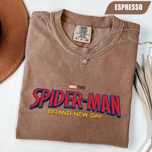 Może przedstawiać: Koszulka w kolorze espresso z logo Spider-Man z Marvel Studios w kolorze czerwonym i niebieskim. Koszulka zawiera ż&oacute;łty napis "BRAND NEW DAY". Srebrny naszyjnik z wisiorkiem jest zarzucony na koszulkę.