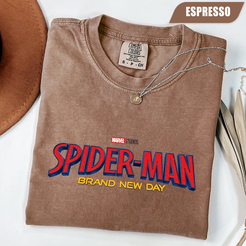 Może przedstawiać: Koszulka w kolorze espresso z logo Spider-Man z Marvel Studios w kolorze czerwonym i niebieskim. Koszulka zawiera ż&oacute;łty napis "BRAND NEW DAY". Srebrny naszyjnik z wisiorkiem jest zarzucony na koszulkę.