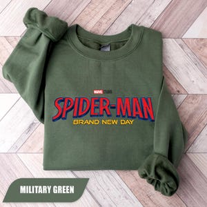 Puede incluir: Una sudadera verde militar con el texto "SPIDER-MAN BRAND NEW DAY" en rojo y azul, con "MARVEL STUDIOS" arriba. La sudadera est&aacute; doblada, con las mangas visibles. Las palabras "MILITARY GREEN" est&aacute;n en una pancarta en la parte inferior.