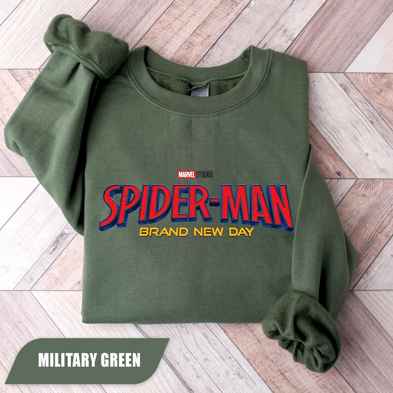 Puede incluir: Una sudadera verde militar con el texto "SPIDER-MAN BRAND NEW DAY" en rojo y azul, con "MARVEL STUDIOS" arriba. La sudadera est&aacute; doblada, con las mangas visibles. Las palabras "MILITARY GREEN" est&aacute;n en una pancarta en la parte inferior.