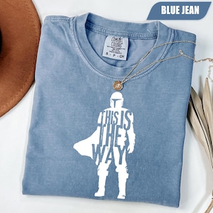 May include: A blue jean colored short-sleeve t-shirt featuring a white graphic of a helmeted character silhouette with the text "THIS IS THE WAY" integrated into the design. The shirt has a crew neckline and a Comfort Colors brand label.