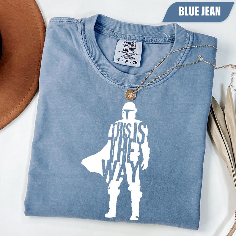 May include: A blue jean colored short-sleeve t-shirt featuring a white graphic of a helmeted character silhouette with the text "THIS IS THE WAY" integrated into the design. The shirt has a crew neckline and a Comfort Colors brand label.