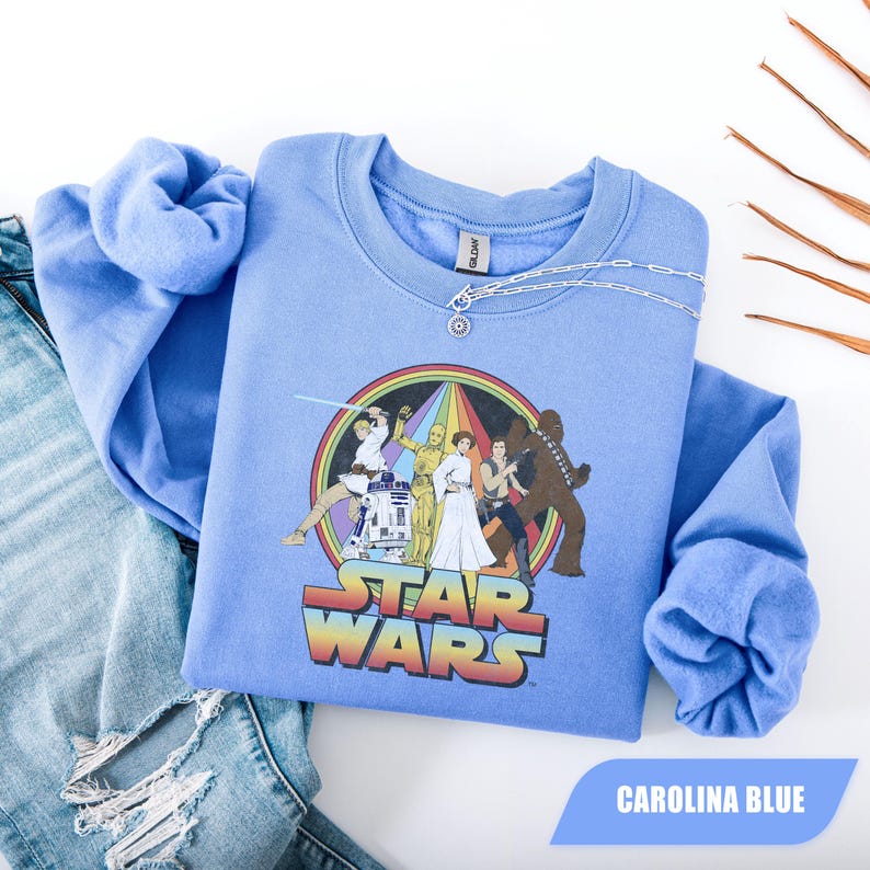 May include: Carolina blue crew neck sweatshirt featuring a Star Wars graphic with characters and the text "STAR WARS" in rainbow colors. A silver chain necklace is draped on the shirt. The sweatshirt is paired with blue jeans.