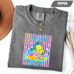 May include: Pepper-colored t-shirt featuring a graphic of a yellow and blue fish with the text "MAKE A SPLASH". The design includes pink stripes, a pink flower, and a seashell. A silver necklace with a pendant is draped across the shirt.