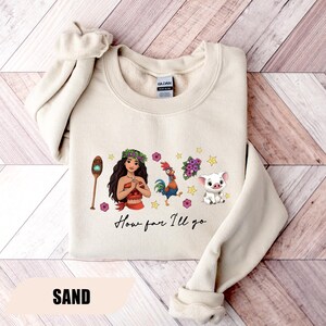 Pu&ograve; includere: Felpa color sabbia con una grafica di una donna polinesiana, un gallo, un maiale e altri elementi. Il testo "How far I'll go" &egrave; scritto in un carattere script. La parola "SAND" &egrave; stampata in basso.