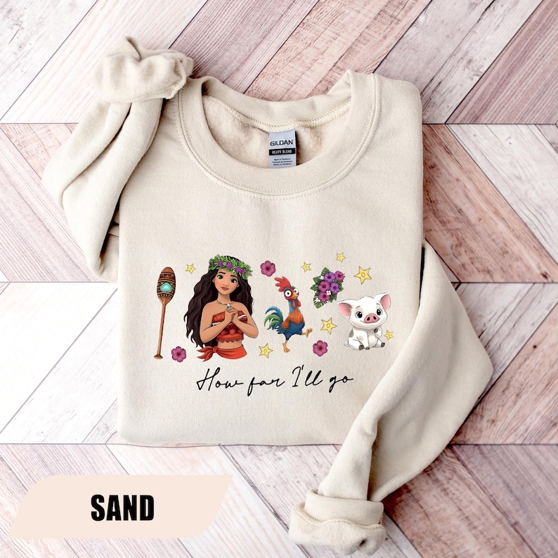 Pu&ograve; includere: Felpa color sabbia con una grafica di una donna polinesiana, un gallo, un maiale e altri elementi. Il testo "How far I'll go" &egrave; scritto in un carattere script. La parola "SAND" &egrave; stampata in basso.