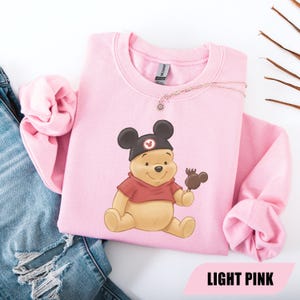 Pu&ograve; includere: Una felpa rosa chiaro con un'illustrazione di Winnie the Pooh con un cappello nero con orecchie di Topolino e che tiene in mano una leccornia al cioccolato. Il testo "LIGHT PINK" &egrave; in basso. Una collana d'oro &egrave; sulla felpa.