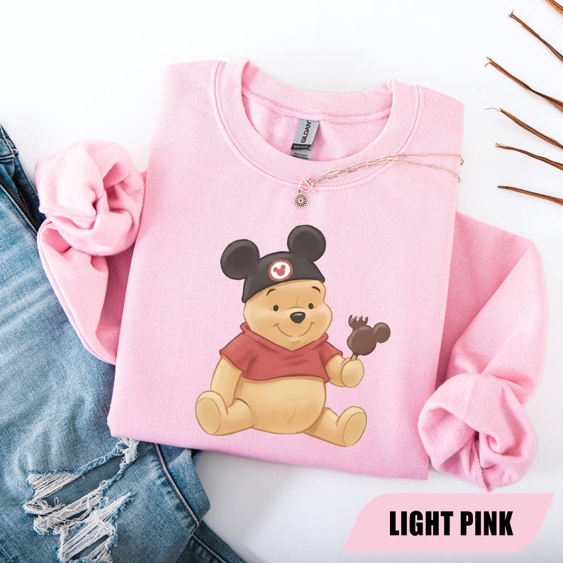 Pu&ograve; includere: Una felpa rosa chiaro con un'illustrazione di Winnie the Pooh con un cappello nero con orecchie di Topolino e che tiene in mano una leccornia al cioccolato. Il testo "LIGHT PINK" &egrave; in basso. Una collana d'oro &egrave; sulla felpa.