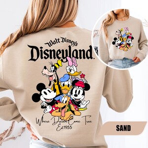 May include: A sand-colored crewneck sweatshirt featuring a large graphic on the back with the text Walt Disney's Disneyland Where Dreams Come True Est 1955 and illustrations of Mickey Mouse, Minnie Mouse, Donald Duck, Daisy Duck, Goofy, and Pluto.