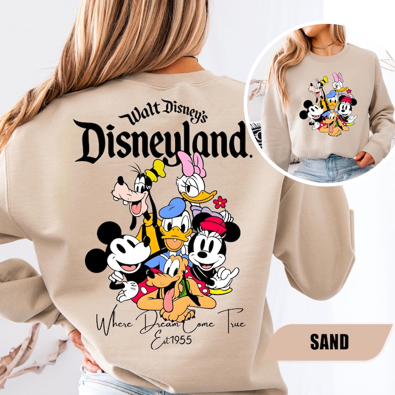 May include: A sand-colored crewneck sweatshirt featuring a large graphic on the back with the text Walt Disney's Disneyland Where Dreams Come True Est 1955 and illustrations of Mickey Mouse, Minnie Mouse, Donald Duck, Daisy Duck, Goofy, and Pluto.