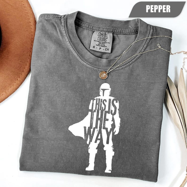 May include: A charcoal gray short-sleeve t-shirt with a white graphic print of a helmeted figure in armor. The text "THIS IS THE WAY" is integrated into the figure's silhouette. The shirt features a Comfort Colors brand label at the neck.