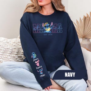 May include: A navy blue crewneck sweatshirt featuring a graphic of the character Stitch wearing a flower crown above the text MAMA and EST. 2026. The sleeve displays three small Stitch characters with the names James, Sophia, and Mike printed below.