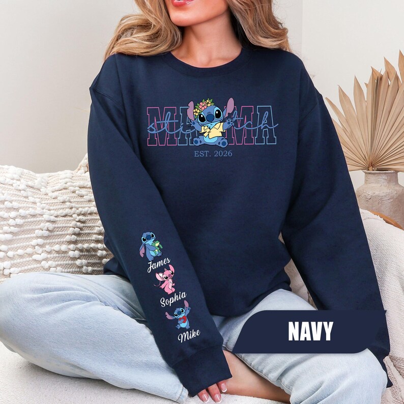 May include: A navy blue crewneck sweatshirt featuring a graphic of the character Stitch wearing a flower crown above the text MAMA and EST. 2026. The sleeve displays three small Stitch characters with the names James, Sophia, and Mike printed below.
