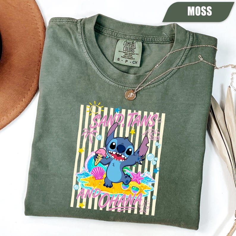 Op de afbeelding: Een mosgroen T-shirt met korte mouwen en een afbeelding van het personage Stitch met een ijshoorntje. Het ontwerp bevat de tekst SAND, TANS AND OHANA, omringd door strandelementen zoals schelpen, sterren en golven.