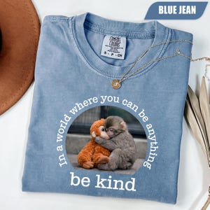 May include: A blue jean coloured t-shirt featuring a circular graphic of a grey monkey hugging an orange stuffed monkey. The text surrounding the image reads, In a world where you can be anything, be kind. The shirt has a crew neckline and a tag.