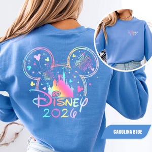 Puede incluir: Sudadera azul Carolina con un dise&ntilde;o colorido de Disney. La parte trasera muestra una gran cabeza de Mickey Mouse con un castillo, fuegos artificiales y el texto "Disney 2026". Una versi&oacute;n m&aacute;s peque&ntilde;a del dise&ntilde;o est&aacute; en el frente. Estilo casual y c&oacute;modo.