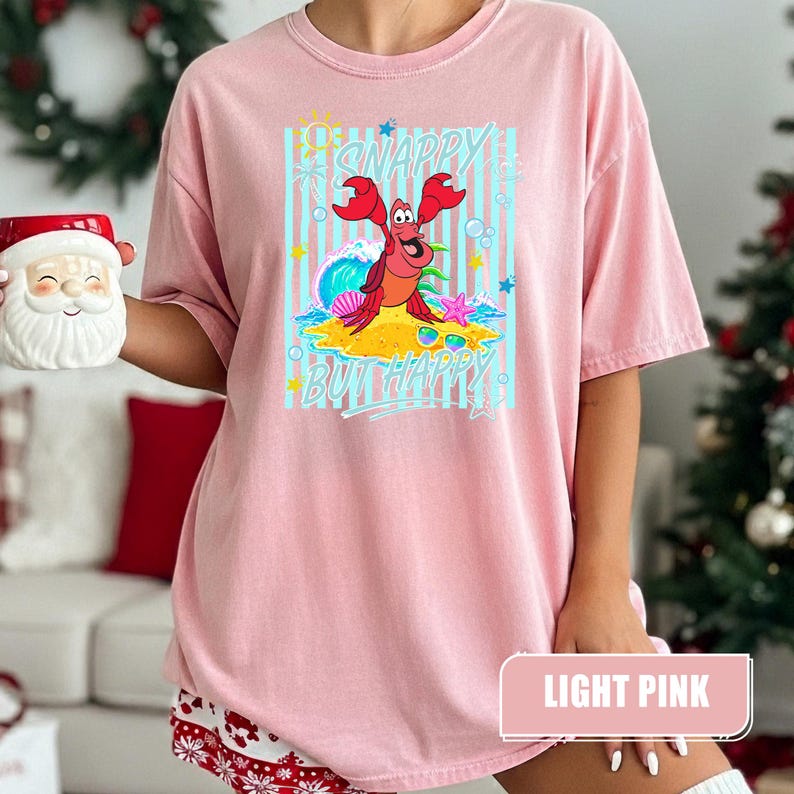 May include: Light pink t-shirt featuring a cartoon crab on a beach with the text "Snappy But Happy". The crab is red with a smiling face. The background has blue stripes and a wave.