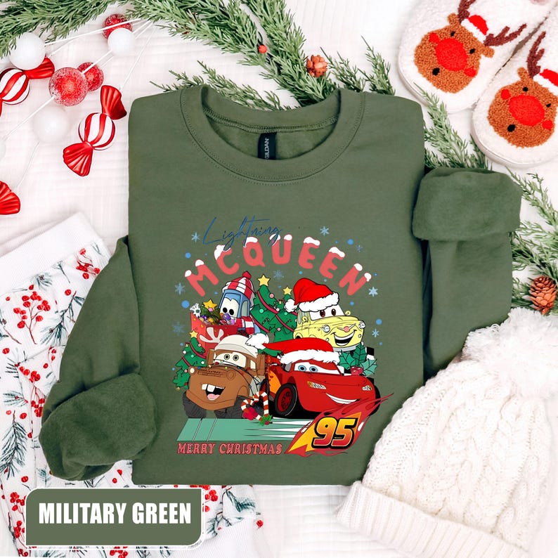 May include: Military green crewneck sweatshirt featuring a graphic of Lightning McQueen and other Cars characters wearing Santa hats and Christmas decorations. The text "Lightning McQueen" and "Merry Christmas" are also displayed.
