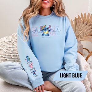 May include: A light blue crewneck sweatshirt featuring a graphic of the character Stitch wearing a flower crown and yellow shirt. The text reads MAMA and EST. 2026. The sleeve displays three small character figures with the names James, Sophia, and Mike.
