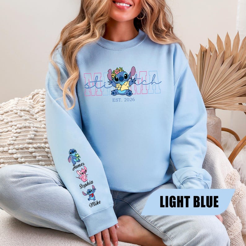 May include: A light blue crewneck sweatshirt featuring a graphic of the character Stitch wearing a flower crown and yellow shirt. The text reads MAMA and EST. 2026. The sleeve displays three small character figures with the names James, Sophia, and Mike.