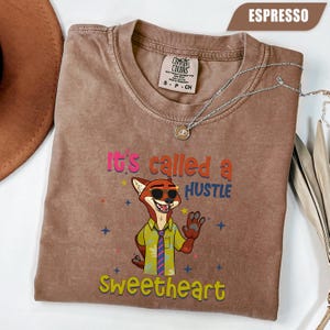 K&ouml;nnte beinhalten: Espressofarbenes T-Shirt mit einem Cartoon-Fuchs, der eine Sonnenbrille und eine Krawatte tr&auml;gt. Auf dem Shirt steht der Text "It's called a HUSTLE Sweetheart" in bunten Buchstaben. Eine silberne Halskette mit Anh&auml;nger liegt auf dem Shirt.
