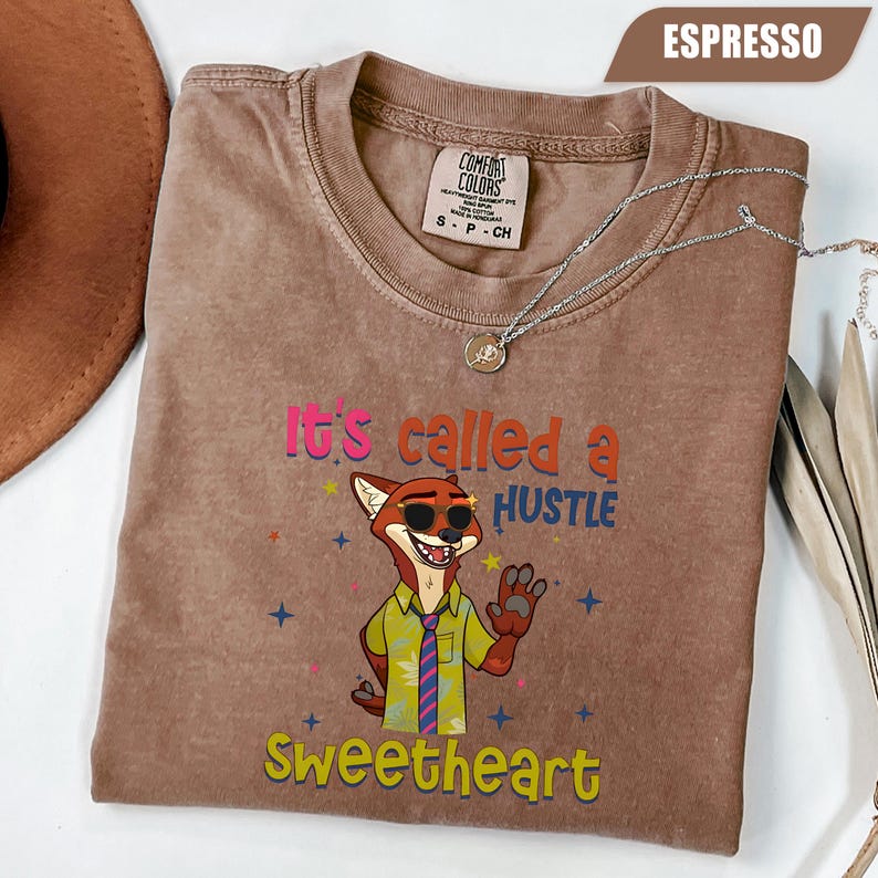 K&ouml;nnte beinhalten: Espressofarbenes T-Shirt mit einem Cartoon-Fuchs, der eine Sonnenbrille und eine Krawatte tr&auml;gt. Auf dem Shirt steht der Text "It's called a HUSTLE Sweetheart" in bunten Buchstaben. Eine silberne Halskette mit Anh&auml;nger liegt auf dem Shirt.