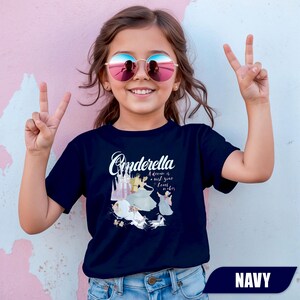 Peut inclure: T-shirt bleu marine avec le mot "Cinderella" en &eacute;criture cursive, et une illustration de Cendrillon et son ch&acirc;teau. Le texte "A dream is a wish your heart makes" est &eacute;galement inclus. V&ecirc;tement pour enfants.