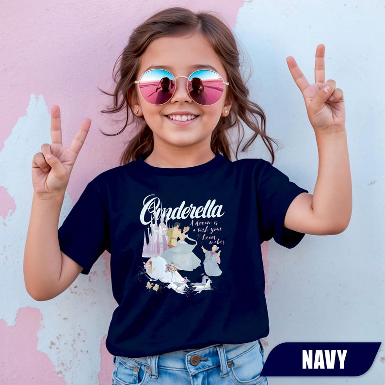 Peut inclure: T-shirt bleu marine avec le mot "Cinderella" en &eacute;criture cursive, et une illustration de Cendrillon et son ch&acirc;teau. Le texte "A dream is a wish your heart makes" est &eacute;galement inclus. V&ecirc;tement pour enfants.