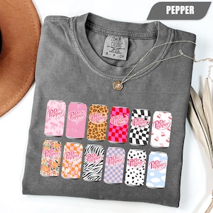 May include: A charcoal gray short-sleeve t-shirt featuring a graphic print of twelve soda cans, each with a unique pattern including leopard print, checkerboard, zebra stripes, and floral designs, all displaying the Dr Pepper logo in pink.