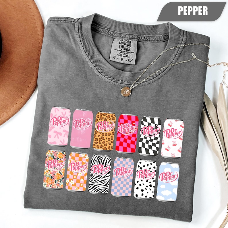 May include: A charcoal gray short-sleeve t-shirt featuring a graphic print of twelve soda cans, each with a unique pattern including leopard print, checkerboard, zebra stripes, and floral designs, all displaying the Dr Pepper logo in pink.