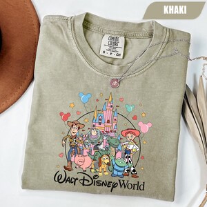 Puede incluir: Camiseta caqui con un gr&aacute;fico colorido de personajes de Toy Story frente a un castillo. El texto "Walt Disney World" est&aacute; impreso debajo. Un collar plateado est&aacute; sobre la camiseta. Talla S-P-CH.