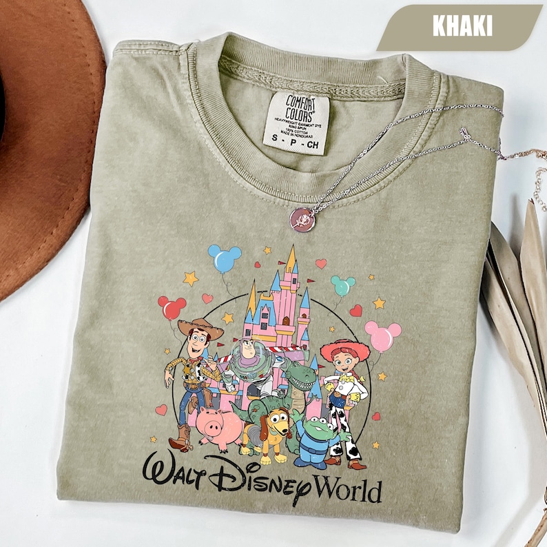 Puede incluir: Camiseta caqui con un gr&aacute;fico colorido de personajes de Toy Story frente a un castillo. El texto "Walt Disney World" est&aacute; impreso debajo. Un collar plateado est&aacute; sobre la camiseta. Talla S-P-CH.
