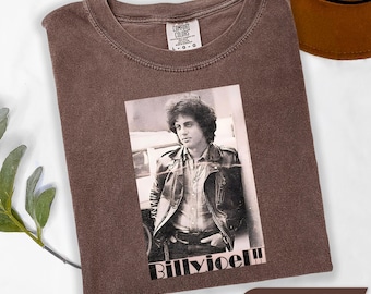 May include: Espresso-colored t-shirt featuring a black and white image of Billy Joel. The image shows Billy Joel in a leather jacket. The text "Billy Joel" is printed below the image. The shirt is made by Comfort Colors.