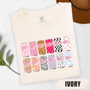 May include: An ivory t-shirt featuring a graphic print of twelve Dr. Pepper soda cans arranged in two rows. Each can displays a unique pattern, including leopard print, zebra stripes, checkerboard, floral, cow print, clouds, bows, and cherries.