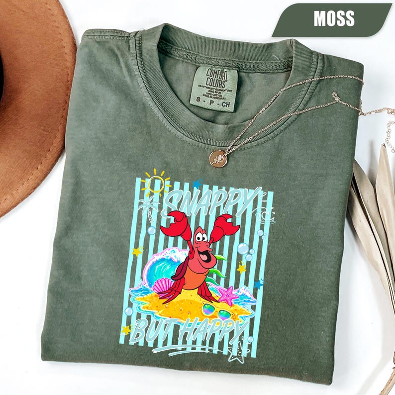 May include: Moss green t-shirt with a graphic of a red crab on a sandy beach with the text "#SNAPPY BUT HAPPY". The shirt has a crew neck and short sleeves. A gold necklace is draped across the shirt.
