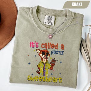 K&ouml;nnte beinhalten: Khaki-farbenes T-Shirt mit dem Text "It's called a HUSTLE Sweetheart" und einem Cartoon-Fuchs mit Sonnenbrille. Das Shirt hat einen Rundhalsausschnitt und kurze &Auml;rmel. Eine silberne Halskette mit Anh&auml;nger befindet sich auf dem Shirt.