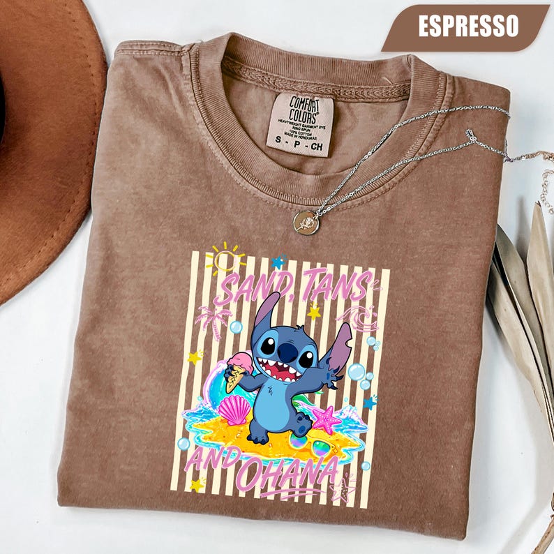 Op de afbeelding: Een espresso-kleurig t-shirt met een afbeelding van het personage Stitch die een ijshoorntje vasthoudt op een strand. Het ontwerp bevat de tekst "SAND, TANS AND OHANA" in het roze, omringd door gele zonnen, sterren en blauwe bubbels.
