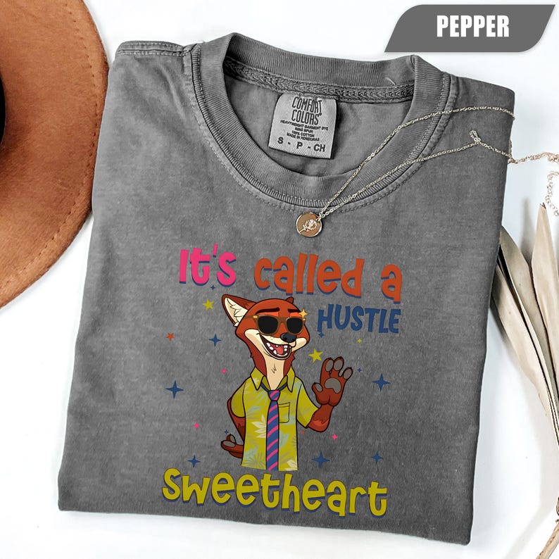 K&ouml;nnte beinhalten: Ein graues Comfort Colors T-Shirt mit dem Schriftzug "It's called a HUSTLE Sweetheart" und einem Cartoon-Fuchs mit Sonnenbrille. Das Shirt hat einen Rundhalsausschnitt und kurze &Auml;rmel. Eine silberne Halskette mit Anh&auml;nger ist auf dem Shirt drapiert.