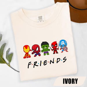 May include: Ivory-colored t-shirt featuring a graphic of cartoon superheroes, including Iron Man, Spider-Man, Hulk, Deadpool, and Captain America. The word "FRIENDS" is printed below the characters in a familiar font. The shirt is labeled "IVORY".
