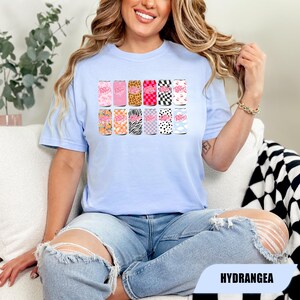 May include: A light blue crew neck t-shirt featuring a graphic print of twelve soda cans arranged in two rows. Each can displays the Dr. Pepper logo and a unique pattern, including leopard print, checkerboard, zebra stripes, floral, and cow print designs.
