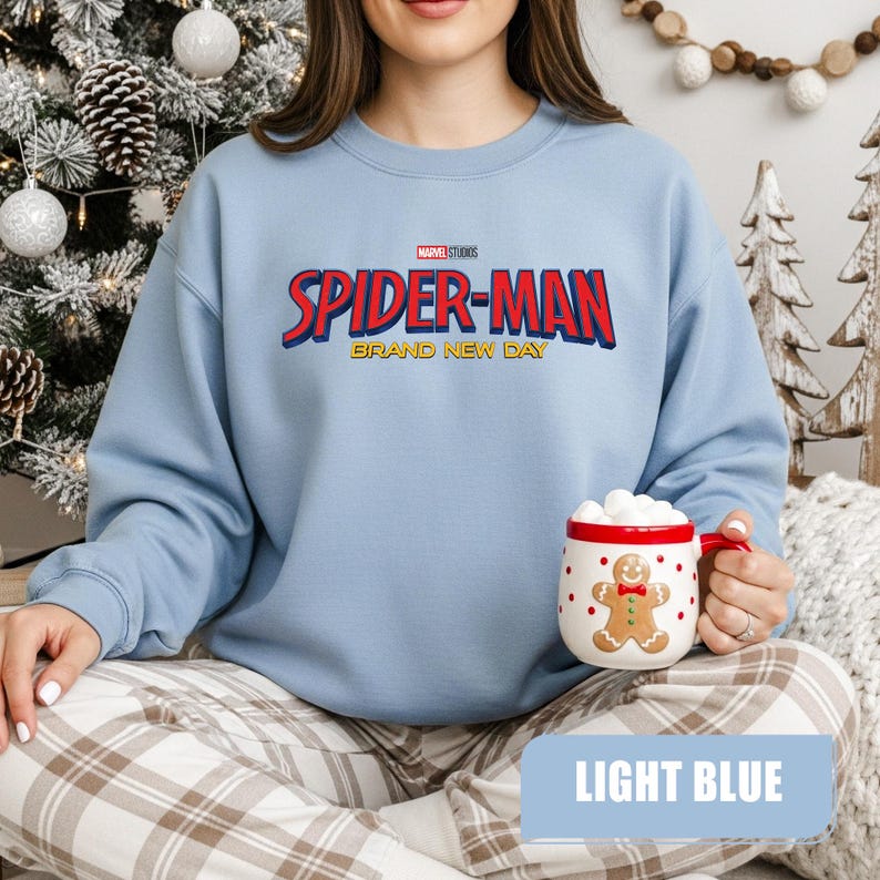 Puede incluir: Sudadera azul claro con el texto "SPIDER-MAN BRAND NEW DAY" en rojo y azul. La sudadera se combina con pantalones de cuadros y una taza de hombre de jengibre con detalles rojos.
