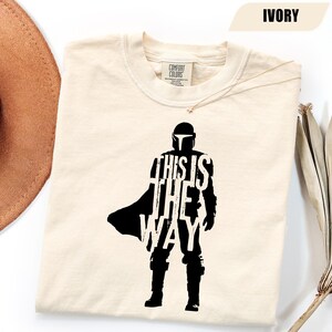 May include: An ivory-colored short-sleeve t-shirt featuring a black silhouette graphic of a helmeted character in armor. The text "THIS IS THE WAY" is printed in white across the center of the figure. The shirt has a Comfort Colors brand label.